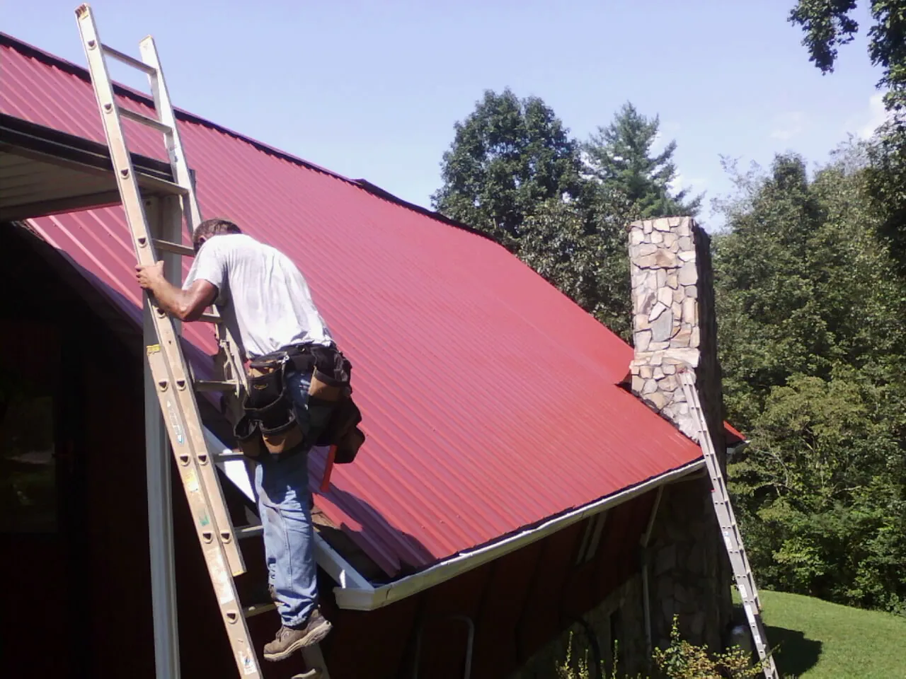 Roofer on a ladder beside a red metal roof and stone chimney in Petersburg
